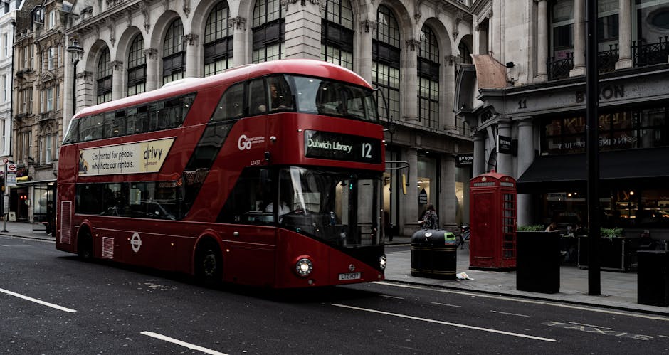 A red double-decker bus parked on a city street in front of historic multi-storey buildings with large windows and classical architectural details. The bus displays the route number 12 and a digital sign indicating it is heading to Dulwich Library. Nearby, there is a traditional red telephone booth positioned on the sidewalk, alongside a black and yellow trash bin. The street surface is dark asphalt, with white lane markings, and the scene is illuminated by natural daylight, highlighting the textures of the bus's shiny exterior, the stone facades, and the smooth surfaces of the street fixtures. This urban environment reflects a typical scene where private transportation and public services coexist, relevant to the context of alternative waste handling and rubbish removal, exemplifying how waste disposal can be an integral part of city management. Waste Disposal Dulwich may often assist with on-site clearance of rubbish, complementing public refuse collection services in busy city areas with historic architecture and street furniture.
