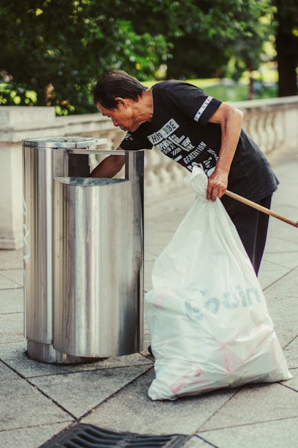 A middle-aged man with dark hair and a black T-shirt is seen outdoors, bending over a metal recycling bin and placing a white plastic bag filled with rubbish into it. The bin is cylindrical, made of shiny stainless steel, and has a large opening at the top. The scene takes place on a paved sidewalk or pathway, with a concrete boundary wall and lush green foliage in the background indicating a park or public space setting. The man uses his right hand to hold the bag open while leaning slightly forward to ensure the rubbish is properly deposited into the bin. The overall atmosphere suggests a routine cleaning or waste disposal activity as part of a community or public waste management effort. The environment is well-lit with natural sunlight, and the surrounding area appears clean and tidy, highlighting the importance of responsible waste disposal and the role of private rubbish collection services like Waste Disposal Dulwich in maintaining area cleanliness and supporting alternative waste handling options.
