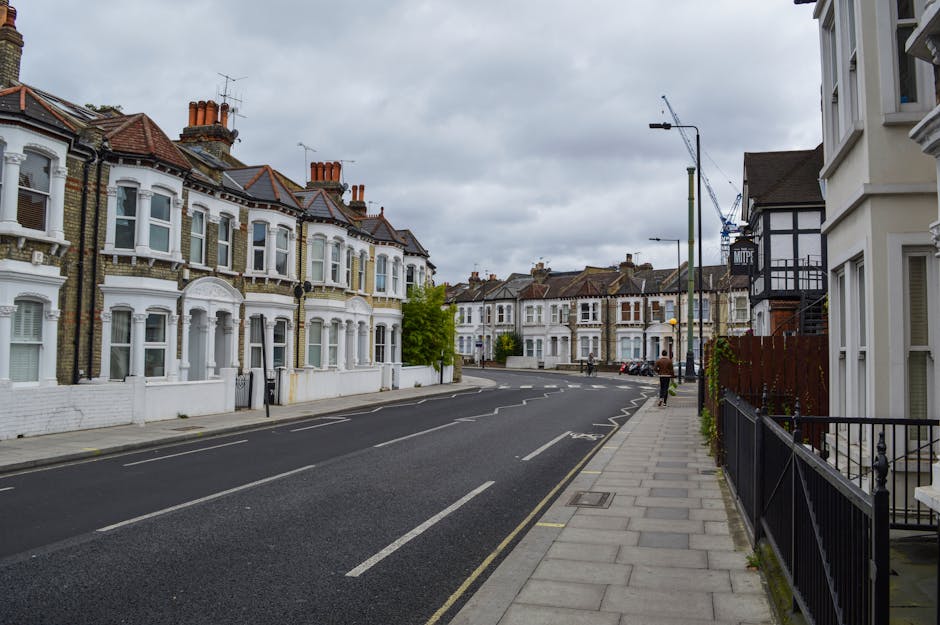 A street scene in a residential neighbourhood with a row of Victorian-style terraced houses on the left side, featuring white and brick facades, bay windows, and chimneys on sloped tiled roofs. The pavement on the right is lined with metal railings and features a few small bushes along the frontage of the houses. The asphalt road has clear white lane markings and curves gently to the right in the distance. Overhead, there are several tall streetlights with simple fixtures, and the sky is overcast with grey clouds, creating diffuse lighting. A person is walking along the sidewalk, and further along the street, there are parked cars in designated bays. In the background, construction cranes are visible above the rooftops, indicating ongoing building or renovation work. The environment suggests a typical urban residential area, with no visible rubbish or waste, consistent with a professional rubbish removal service in the area, such as Waste Disposal Dulwich, providing ongoing or alternative waste management solutions.