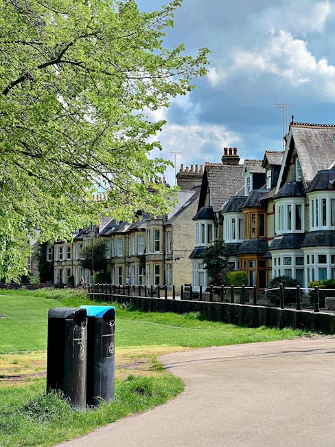 A row of multi-story Victorian-style terraced houses with ornate metal and woodwork details on their facades, large front bay windows, and small balconies. The buildings are constructed from brick with painted detailing in white, and have pitched roofs with gables. The houses are situated on a slightly elevated street level, with brick retaining walls and small landscaped front gardens featuring shrubs and plants. A series of black wheelie bins are lined up along the pavement in front of the properties, indicating waste collection. The background shows a clear sky with a few wispy clouds, and the area appears to be in a residential urban setting, possibly prepared for or reflecting private waste management services, consistent with alternative rubbish removal options like those offered by Waste Disposal Dulwich.