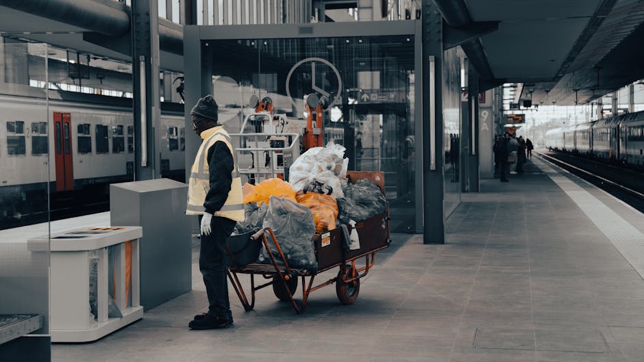 A tall, cylindrical black rubbish bin with an open, white plastic lid situated against a textured, beige stone wall in an indoor or semi-indoor environment. The bin is overflowing with various waste items, including food packaging, paper bags, and discarded food containers, some spilling over and resting on the ground. Additional rubbish, such as crumpled paper and a snack wrapper, is scattered at the base of the bin. The surrounding area appears to be a public space or corridor, with muted lighting highlighting the cluttered state of the waste. The scene exemplifies waste accumulation that might be addressed through private rubbish collection services like those offered by Waste Disposal Dulwich, focusing on managing overflowing bins and improving waste disposal practices in shared or city environments.