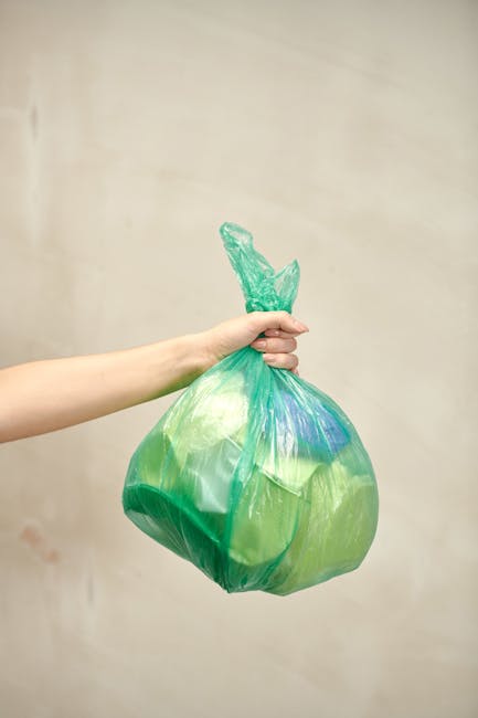 A person's hand and forearm are visible, holding a green plastic rubbish bag by its tied top. The bag appears semi-full with visible bulges indicating contents inside, and it has a smooth, slightly glossy surface. The background is a plain, light-colored wall, creating a neutral environment for the image. The scene suggests an act of waste disposal or rubbish collection, potentially related to private waste handling services such as those offered by Waste Disposal Dulwich. The bag's size and the way it is being held imply it is intended for removal, fitting within the context of rubbish clearance. There are no additional objects or environmental details visible, keeping the focus on the waste bag and the act of rubbish disposal, which aligns with themes of independent collection or alternative waste management methods.