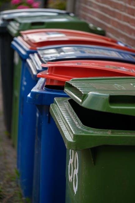 A row of large, outdoor trash bins arranged along a narrow pavement adjacent to a brick wall. The bins are made of durable plastic with textured lids, each painted in distinct colors to indicate different waste types; the first bin in the foreground is green with a hinged lid, followed by other bins in blue, red, and green, all featuring slotted lids for ventilation. The bins are positioned closely together, with some lids slightly open, and are set on a concrete or paved surface next to a residential building or boundary wall. The environment appears to be an urban or suburban street scene during daylight, with natural lighting highlighting the different colors and textures of the bins, subtly emphasizing the importance of proper waste segregation and disposal. This image aligns with private waste collection services such as those provided by Waste Disposal Dulwich, illustrating the common sight of municipal or private rubbish bins in the context of local rubbish removal and alternative waste handling options.