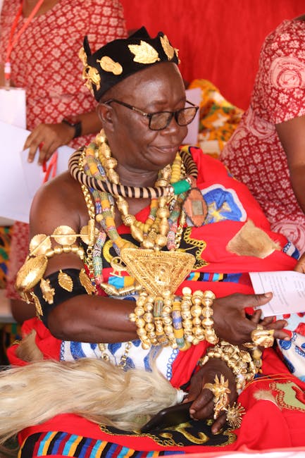 A distinguished woman dressed in traditional African attire adorned with elaborate gold jewelry, including multiple layered necklaces, large bracelets, and a decorative headpiece featuring gold accents and intricate patterns. She is seated indoors against a vibrant red fabric background, with other individuals wearing similarly colorful textiles partially visible around her. The woman holds a piece of paper in her hand, and her expression appears focused or contemplative. Her outfit includes bright red, black, and multicolored elements, with textured fabrics and ornate embellishments, reflecting cultural heritage and ceremonial attire often associated with significant cultural or social gatherings. The setting suggests a formal or celebratory occasion, with a richly decorated environment that emphasizes traditional craftsmanship and opulence, aligning with themes of cultural pride and heritage celebration. This scene highlights the significance of traditional dress and adornment in cultural events, and it subtly connects to the broader context of community, identity, or cultural heritage preservation.